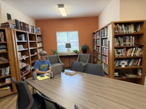 A congregation member uses the New Spirit Lutheran Church library as a remote workspace, sitting at a large table surrounded by bookshelves and comfortable seating.